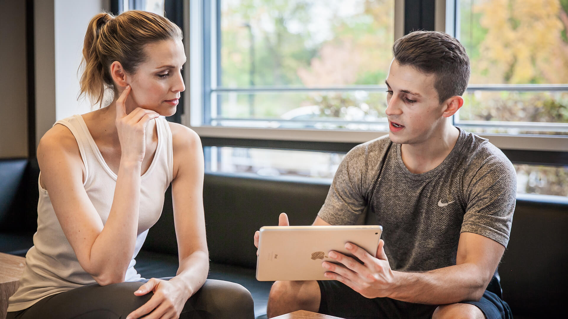 Ein Fitnesstrainer bespricht mit einer Kundin auf einem Tablet Trainingspläne oder Fortschritte. Beide sitzen entspannt auf einer Bank im Studio.